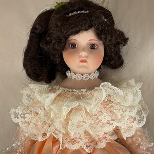 May include: A porcelain doll with brown curly hair and brown eyes. The doll wears a peach-colored satin dress with a lace collar and a jeweled choker. A small floral hair accessory is visible.