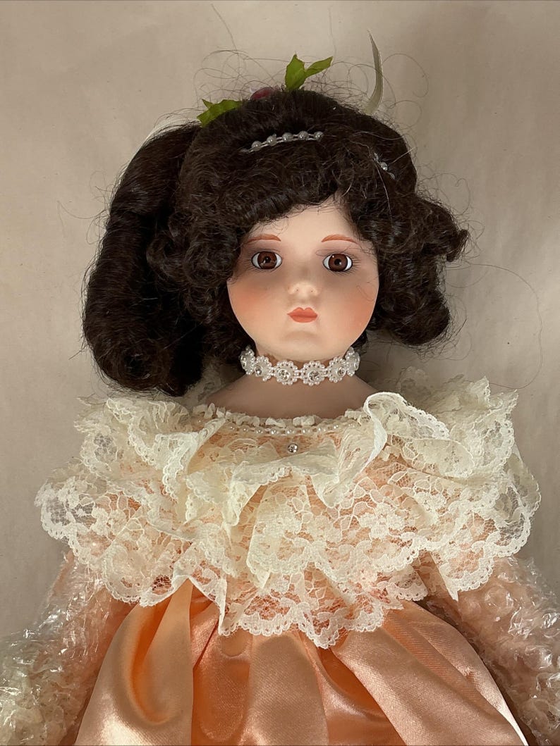 May include: A porcelain doll with brown curly hair and brown eyes. The doll wears a peach-colored satin dress with a lace collar and a jeweled choker. A small floral hair accessory is visible.