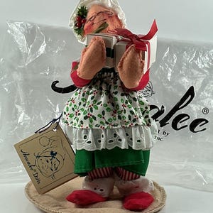 Pu&ograve; includere: Una bambola Annalee fatta a mano, una figura a tema natalizio, che tiene due piccoli regali avvolti legati con un nastro rosso. La bambola indossa un cappello bianco con agrifoglio, un vestito a fantasia verde e bianco e scarpe rosse. Un'etichetta recita "Annalee Dolls".
