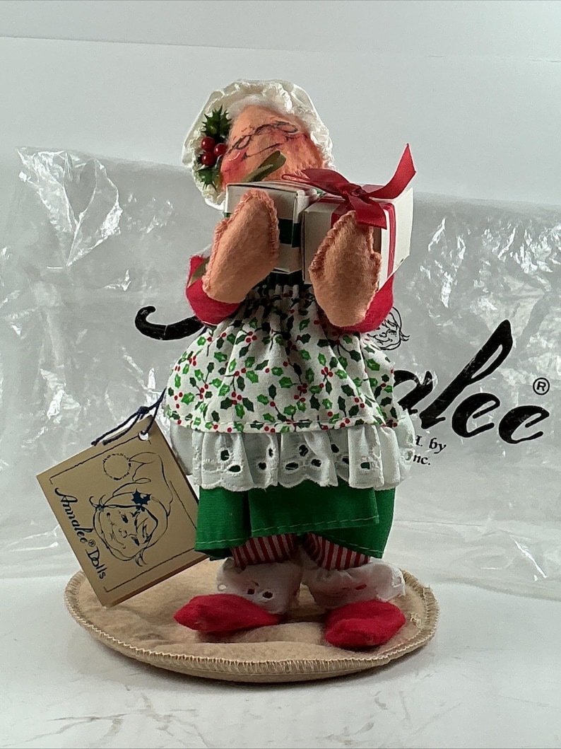 Pu&ograve; includere: Una bambola Annalee fatta a mano, una figura a tema natalizio, che tiene due piccoli regali avvolti legati con un nastro rosso. La bambola indossa un cappello bianco con agrifoglio, un vestito a fantasia verde e bianco e scarpe rosse. Un'etichetta recita "Annalee Dolls".