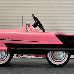 May include: A pink and black toy car with the word "Kidillac" on the side. The car has a steering wheel and a chrome bumper.