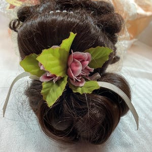 May include: A dark brown wig with a bun hairstyle, adorned with a pink rose and green leaves. A light gray ribbon is tied around the bun. The wig is styled with curls and waves, and is likely for costume or theatrical use.