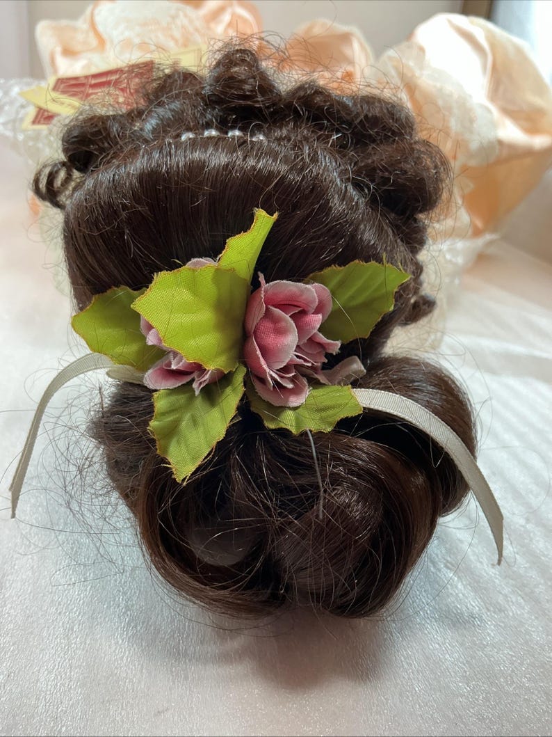 May include: A dark brown wig with a bun hairstyle, adorned with a pink rose and green leaves. A light gray ribbon is tied around the bun. The wig is styled with curls and waves, and is likely for costume or theatrical use.