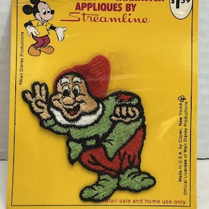 May include: A Walt Disney Character applique by Streamline featuring an embroidered patch of the dwarf Dopey on a yellow backing card. The patch depicts the character in a red hat, green tunic, and red pants, with text including Walt Disney Productions.