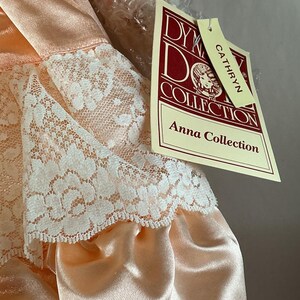 May include: A peach-colored doll dress with white lace trim. The dress is part of the Anna Collection by Dykeman Doll Collection. The tag reads "Cathryn". The dress is made of a satin-like material.