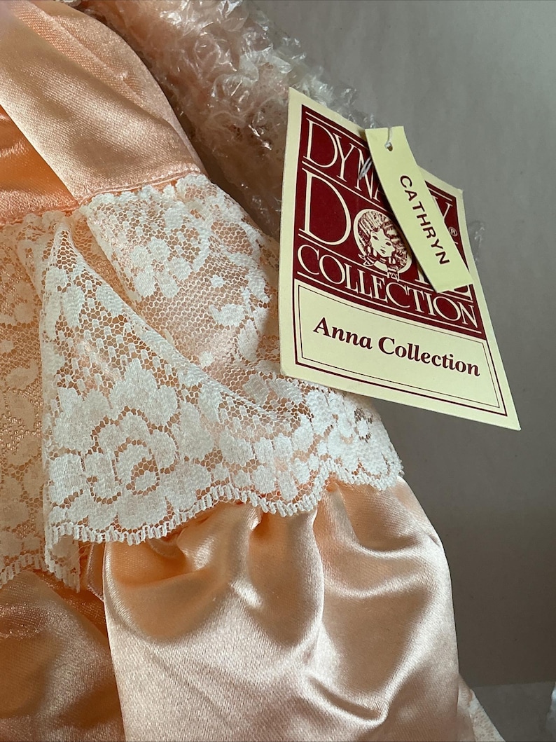 May include: A peach-colored doll dress with white lace trim. The dress is part of the Anna Collection by Dykeman Doll Collection. The tag reads "Cathryn". The dress is made of a satin-like material.