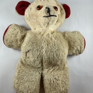 May include: A vintage teddy bear with a cream-colored body and red accents on the ears, hands, and feet. The bear has brown button eyes and a stitched black nose and mouth. The plush toy is in a well-loved condition.