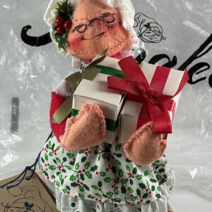 Pu&ograve; includere: Bambola fatta a mano di Mrs. Claus, con un cappello bianco, occhiali e un'espressione sorridente. Indossa un abito rosso e bianco, una gonna a fantasia di agrifoglio verde e bianco e tiene in mano due regali avvolti con nastri rossi e verdi. &Egrave; attaccata un'etichetta con un disegno di bambola.