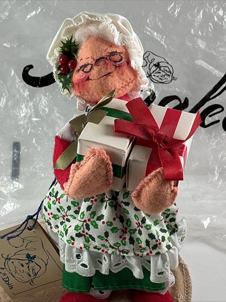 Pu&ograve; includere: Bambola fatta a mano di Mrs. Claus, con un cappello bianco, occhiali e un'espressione sorridente. Indossa un abito rosso e bianco, una gonna a fantasia di agrifoglio verde e bianco e tiene in mano due regali avvolti con nastri rossi e verdi. &Egrave; attaccata un'etichetta con un disegno di bambola.