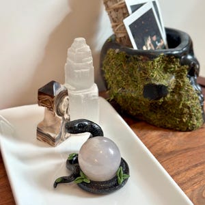 May include: A decorative white rectangular tray holds a black snake figurine with a pink crystal ball. Other items include a selenite tower, a brown and white stone, and a skull-shaped container filled with moss and sage bundles.
