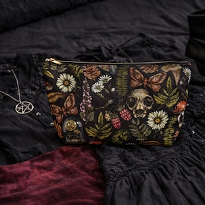 May include: Black zippered cosmetic bag with a botanical print featuring butterflies, flowers, mushrooms, and a skull. The bag has a gold zipper and is set against a dark background.