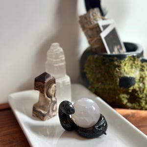 May include: A white rectangular tray holds a black snake figurine coiled around a light pink crystal ball. Other items include a brown and white stone, and a clear crystal tower. A black skull-shaped container with moss and sage bundles is in the background.