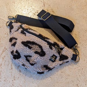 Könnte beinhalten: Eine beige-schwarze Gürteltasche mit Leopardenmuster und einem schwarzen, verstellbaren Riemen. Die Tasche hat einen schwarzen Reißverschluss und ein kleines Etikett mit der Aufschrift "VERS LOOK". Die Tasche ist aus weichem, flauschigem Material.