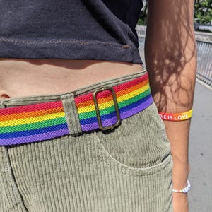 Könnte beinhalten: Ein regenbogenfarbener Stoffgürtel mit einer rechteckigen, bronzefarbenen Metallschnalle, getragen über einer olivgrünen Cordhose. Ein gelb-oranges Armband mit dem Text LOVE IS LOVE ist am Arm sichtbar, zusammen mit einem kleinen Perlenarmband.