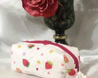 May include: A white quilted cosmetic bag with a strawberry and floral pattern. The bag has a bright pink zipper and a matching fabric pull. The background includes a vase of flowers.