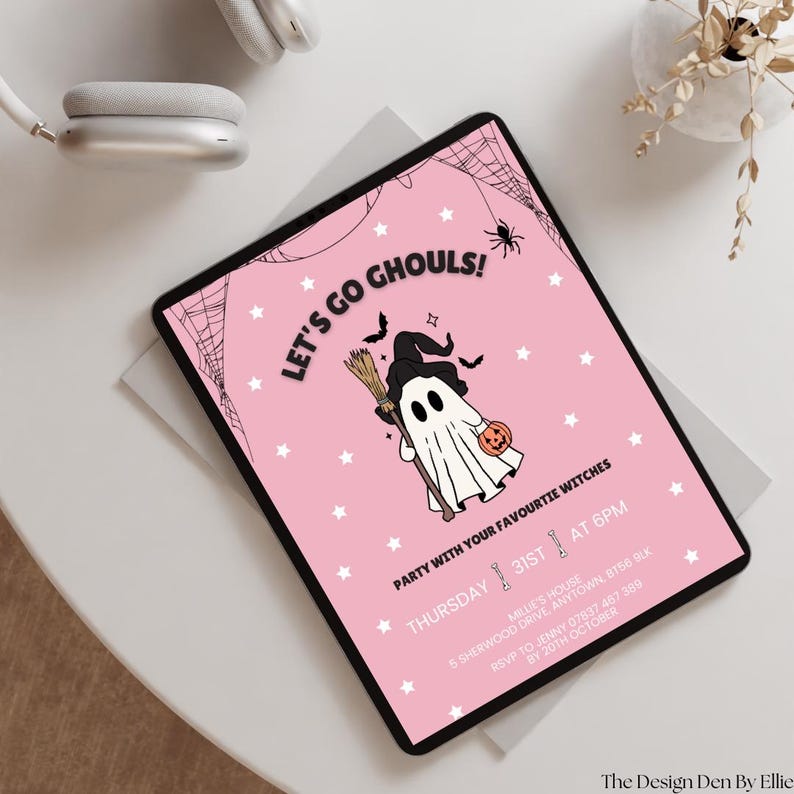 May include: A pink Halloween party invitation on a tablet. The invitation features a cartoon ghost holding a pumpkin and broom, with the text "Let's Go Ghouls!" and event details. A pair of white headphones and a small plant are in the background.