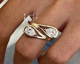 May include: A gold ring with two pear-shaped, clear gemstones. The ring has a unique, open design with a polished finish. The ring is worn on a finger, showcasing its elegant and modern style. Jewelry.