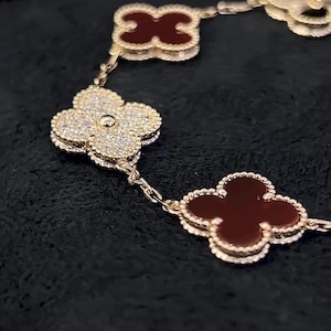 Puede incluir: Una pulsera dorada con abalorios en forma de trébol. Algunos abalorios están incrustados con pequeñas piedras brillantes, mientras que otros tienen un acabado rojo oscuro y brillante. La pulsera se muestra sobre un fondo oscuro.