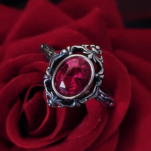 Peut inclure: Une bague en argent avec un motif orné et une grande pierre précieuse rouge sertie au centre. La bague repose sur une rose rouge.
