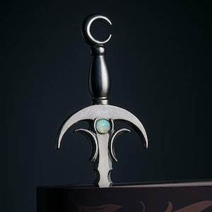 Pode incluir: Um marcador de página prateado com uma pega em forma de lua crescente e uma pedra semelhante à opala. O marcador de página decorativo está colocado sobre um fundo escuro, com um design floral detalhado visível na capa do livro.