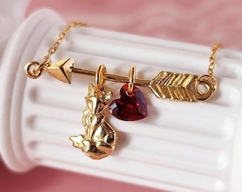 Op de afbeelding: Een gouden ketting met een bedel in de vorm van een pijl met een rood hart en een vosbedel.