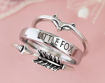 Op de afbeelding: Drie zilveren ringen op elkaar gestapeld. De bovenste ring heeft een klein vosje ontwerp. De middelste ring heeft de woorden "LITTLE FOX" gegraveerd. De onderste ring heeft een pijl ontwerp met een veer.