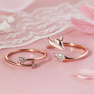 Puede incluir: Dos anillos de oro con diseños delicados. Un anillo presenta una pequeña flecha con un solo diamante, mientras que el otro anillo tiene una cabeza de zorro estilizada con una fila de diamantes.