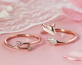 Op de afbeelding: Twee gouden ringen met delicate ontwerpen. Een ring heeft een kleine pijl met een enkele diamant, terwijl de andere ring een gestileerd vossenhoofd heeft met een rij diamanten.
