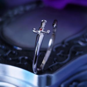 Op de afbeelding: Een zilveren ring met een dolk-ontwerp. De dolk heeft een rode edelsteen aan de bovenkant. De ring ligt op een zilveren doos met een versierd ontwerp.