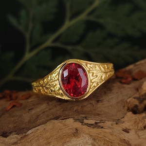 以下が含まれることがあります： 楕円形の赤い宝石が付いた金の指輪。リングバンドには、複雑な葉のデザインが施されています。指輪は木製の表面に置かれ、背景はぼやけた緑色です。