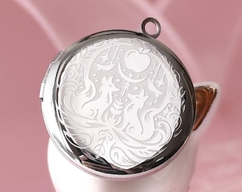 Op de afbeelding: Een zilverkleurige medaillon met een gedetailleerd gegraveerd ontwerp met vossen, een maan en bladeren. De ronde medaillon heeft een donkere rand en een lus voor een ketting. De achtergrond is zachtroze.