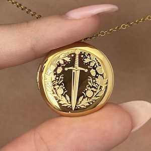 May include: A gold-colored round pendant necklace. The pendant features a sword design in the center, surrounded by a decorative border of leaves and berries. The necklace is held by a person's fingers, showcasing the intricate details of the jewelry.