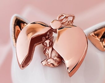 Op de afbeelding: Een roodgouden hartvormige locket hanger met een kleine vos figuur binnenin. De locket is open, onthullend de vos. De hanger staat op een witte keramische katvormige houder met gouden accenten.
