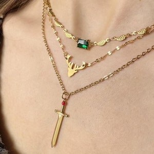 May include: Three gold necklaces with pendants. The top necklace has a green gemstone pendant. The middle necklace has a deer head pendant. The bottom necklace has a sword pendant with a red jewel.