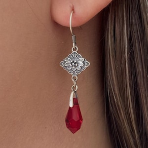 May include: Silver dangle earrings with a red crystal teardrop pendant. The earrings feature a silver filigree design with a floral motif.