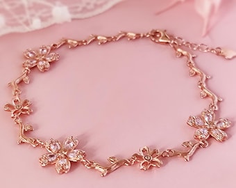 Op de afbeelding: Een delicate armband van roségoud met een bloemenmotief. De armband heeft vijf kleine bloemen met heldere stenen, verbonden door een rankachtige ketting.