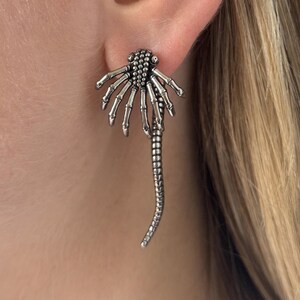 Könnte beinhalten: Ein silberfarbener Ohrring mit einem Skeletthand-Design. Der Ohrring hat eine detaillierte Handfläche und Finger mit einem langen, strukturierten, hängenden Element. Der Ohrring wird an einem Ohr getragen und zeigt sein kompliziertes Design.