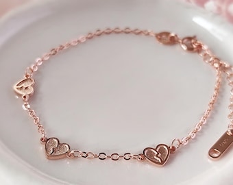 Op de afbeelding: Een delicate roségouden armband met drie hartbedels, waarvan één met een gebroken hart ontwerp. De armband ligt op een wit bord met een zachte roze achtergrond. De armband heeft een klein label met de tekst "S925".