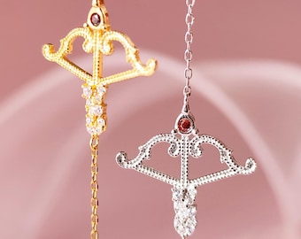 Op de afbeelding: Twee delicate kettingen met boog- en pijlhangers. De bovenste ketting heeft een gouden ketting en een bruine edelsteen in de pijl. De onderste ketting heeft een zilveren ketting en een rode edelsteen in de pijl. Beide hangers zijn versierd met kleine, heldere edelstenen.