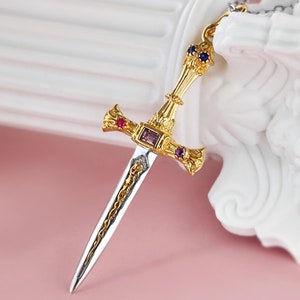 May include: A gold and silver sword pendant necklace with red and blue gemstones. The sword has intricate details and a pointed tip.