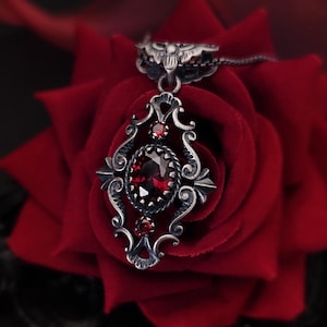 Peut inclure: Un pendentif en argent avec une pierre précieuse rouge dans un design orné détaillé, reposant sur une rose en velours rouge.
