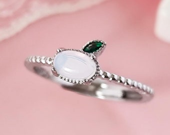 Op de afbeelding: Een zilveren ring met een witte ovale steen en een klein groen steentje erop. De ring ligt op een roze achtergrond.