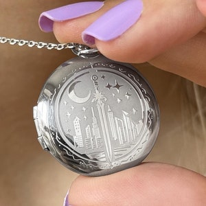 Puede incluir: Collar con dije de plata con un diseño detallado de ciudad, que incluye una luna creciente, estrellas y un edificio alto con una punta puntiaguda. El dije está abierto y el interior es visible.