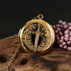 May include: A gold-colored locket necklace with a detailed design. The round locket features a sword and wreath engraving. The chain is also gold. The background is a dark color with a wooden surface.