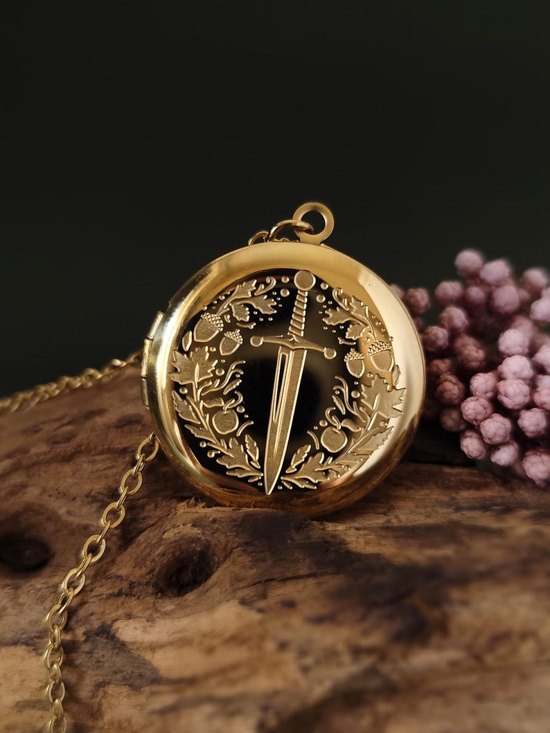 May include: A gold-colored locket necklace with a detailed design. The round locket features a sword and wreath engraving. The chain is also gold. The background is a dark color with a wooden surface.