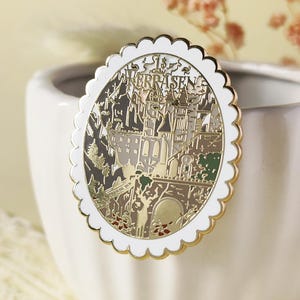 以下が含まれることがあります： スカラップの白い縁と金のトリムが付いた丸いエナメルピン。ピンは、グレー、金、緑の色合いで詳細な都市景観のデザインを特徴とし、上部に「TERRASEN」という単語があります。ピンは白いセラミック容器にセットされています。