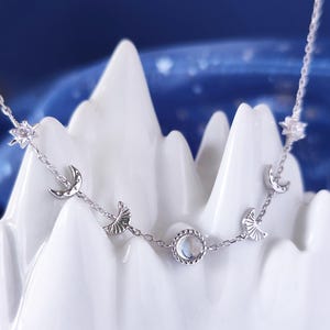 May include: A silver necklace with a moonstone pendant and crescent moon charms. The necklace is on a white background.