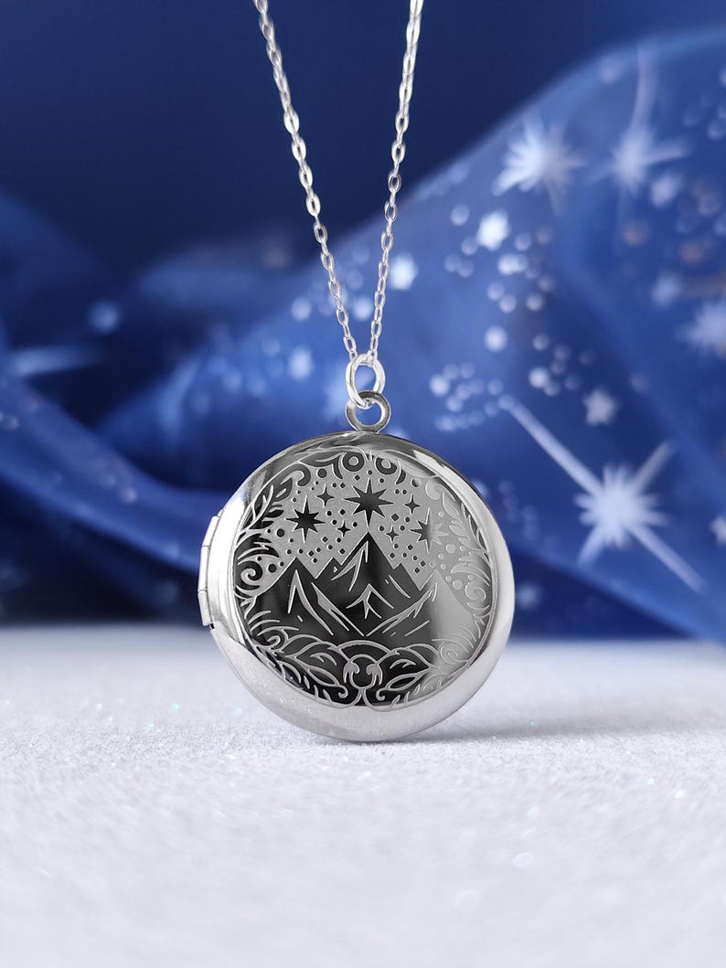 May include: A silver locket necklace with a detailed design of mountains, stars, and a crescent moon. The locket is round and has a hinged opening.