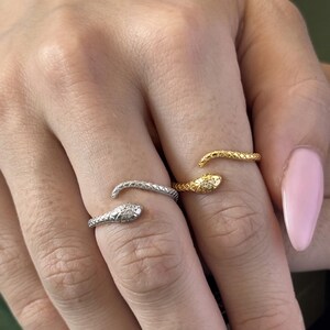 May include: Three snake rings in silver and gold tones are displayed on a hand with pink nail polish. The rings are wrapped around the fingers, with the snake heads facing outward. One ring has a small diamond accent.