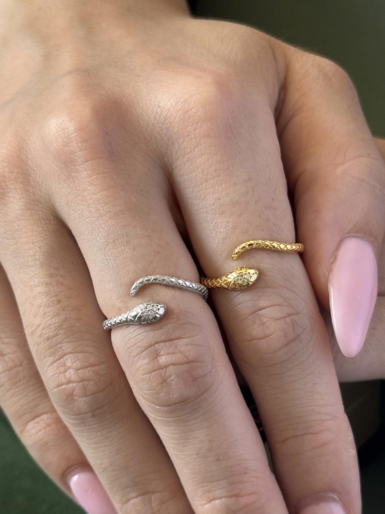 May include: Three snake rings in silver and gold tones are displayed on a hand with pink nail polish. The rings are wrapped around the fingers, with the snake heads facing outward. One ring has a small diamond accent.